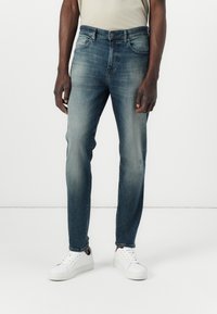 Jeans de mezclilla azul oscuro con corte ajustado, que cuentan con un lavado desvanecido, un diseño de cinco bolsillos y costuras claras. Combinados con zapatillas blancas.