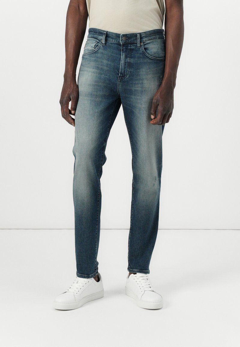 Jeans de mezclilla azul oscuro con corte ajustado, que cuentan con un lavado desvanecido, un diseño de cinco bolsillos y costuras claras. Combinados con zapatillas blancas.