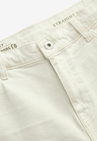 Pantalons beige coupe droite avec une texture lisse, fermeture à bouton en métal et détails de couture. L'étiquette indique "COUPE DROITE" et le nom de la marque est visible.