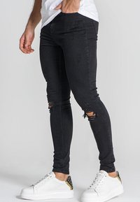 Jeans negros ajustados con desgastes desgarrados en las rodillas, combinados con zapatillas blancas que tienen un acento dorado en la parte trasera. Textura de denim suave.