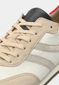 Κοντινό πλάνο μπεζ και λευκού αθλητικού παπουτσιού με κρεμ κορδόνια, μαύρη γλώσσα και υφασμάτινες πλευρικές ρίγες.