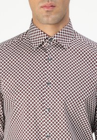 Chemise à boutons en tissu léger, avec une base blanche ornée d'un motif en losanges rouges, un col allemand et des boutons gris.