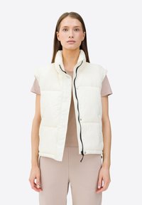 Gilet imbottito crema con colletto alto, bottoni a pressione e dettagli con zip. Design senza maniche con texture trapuntata e accenti neri.