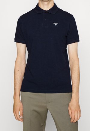 Homme portant un polo bleu marine uni avec le logo Barbour sur la poitrine gauche et un pantalon marron clair, debout devant un fond clair.