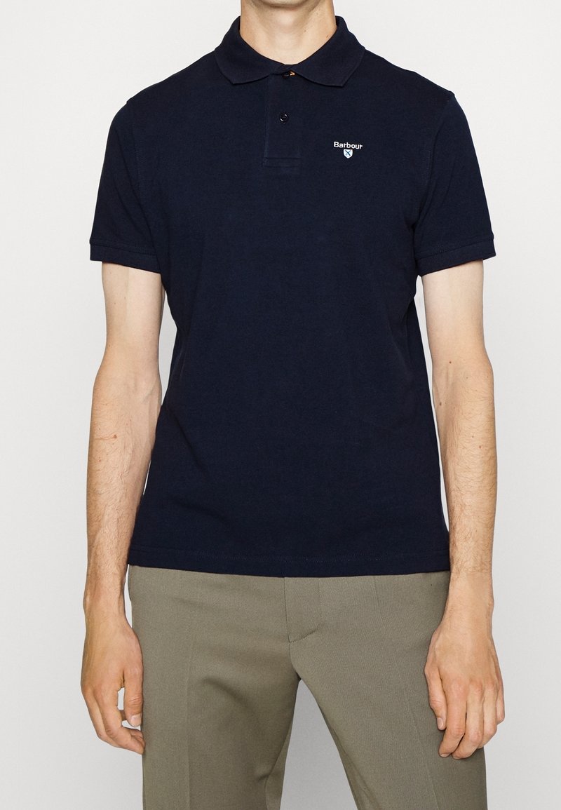 Homme portant un polo bleu marine uni avec le logo Barbour sur la poitrine gauche et un pantalon marron clair, debout devant un fond clair.
