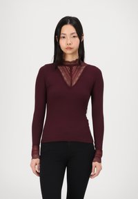 Burgundy pitkähihaisessa paidassa on pitsireunukset kaula-aukossa ja hihoissa, se on muotoiltu ja ribattu. Ei näkyviä logoja tai kuvioita.