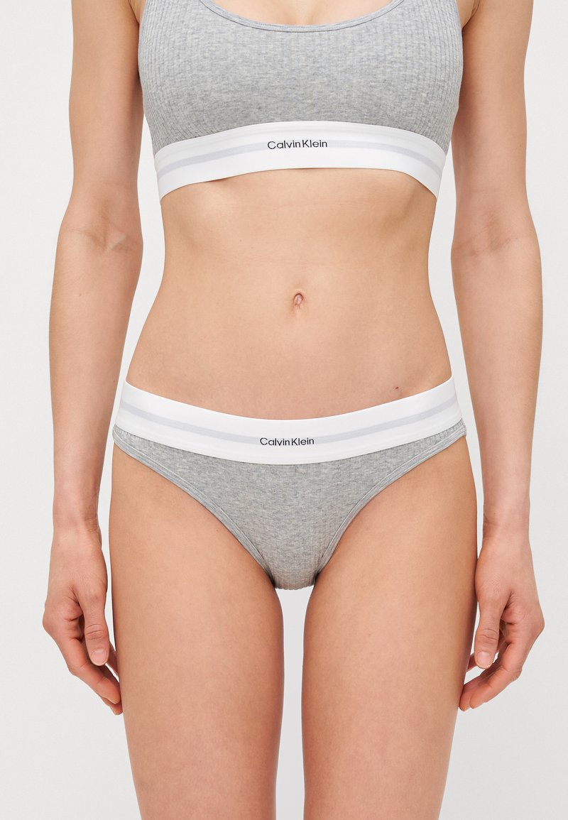 Bralette grigia a coste e mutandine bikini abbinate, caratterizzate da un elastico bianco con il marchio "Calvin Klein" e design minimalista.