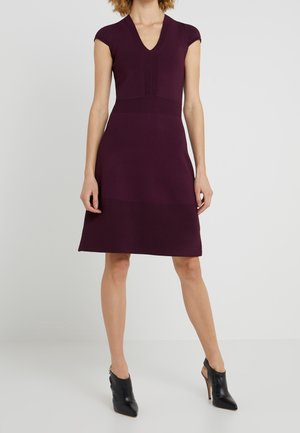 Frau trägt ein knielanges, ärmelloses Pflaumenkleid mit V-Ausschnitt und schwarze, hochhackige Stiefeletten, steht vor unifarbenem Hintergrund.