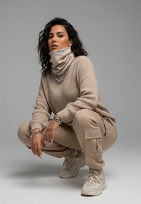 Beige knitted sweater with a high turtleneck, matching beige cargo pants, and light sneakers. Silver chain accents on the neck and wrist.