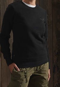 Sudadera negra con cuello redondo, hecha de una tela suave, que presenta un sutil logo en el pecho y puños y dobladillo de canalé texturizados.