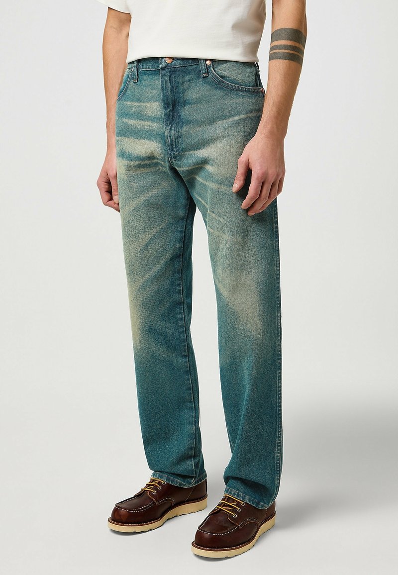 Homme portant un jean bleu délavé à jambes droites, une chemise blanche, des chaussures en cuir marron et un tatouage de brassard noir sur l'avant-bras droit.