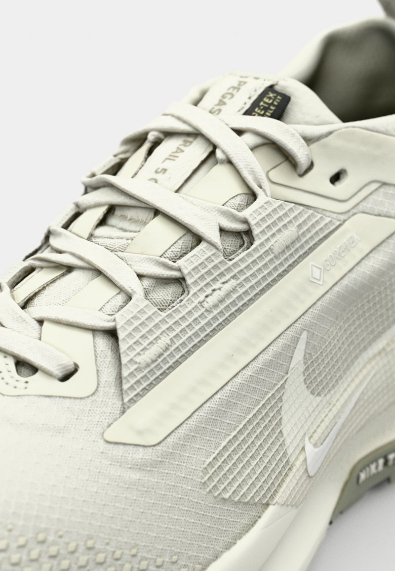 Jasnoszary but do biegania w terenie, z teksturowaną cholewką, gumową podeszwą i detalami z Gore-Tex; sznurowadła i logo podkreślają design.