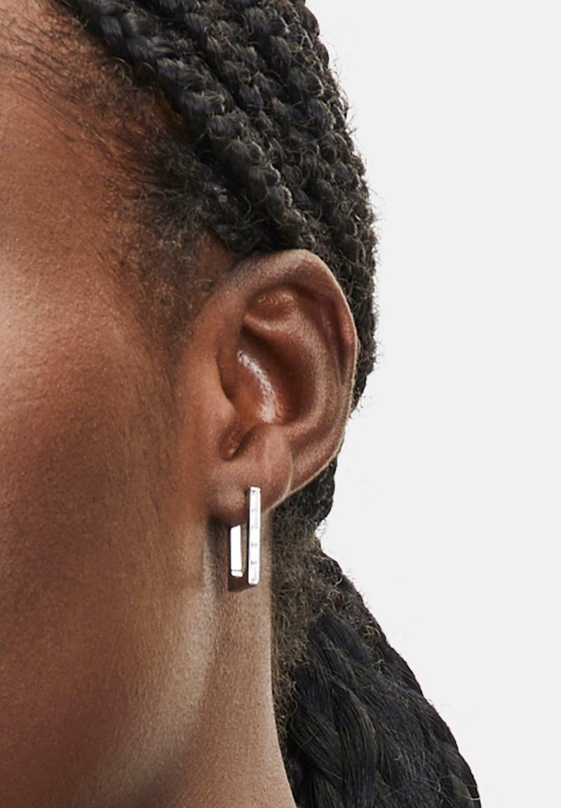 Silberne rechteckige Ohrringe hängen vom Ohr und bieten ein poliertes Finish mit einem matten Silberakzent. Texturiertes, geflochtenes Haar ist sichtbar.