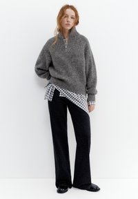 Młoda kobieta stojąca, ubrana w szary sweter na zamek, koszulę w kratę, czarne spodnie o szerokich nogawkach i czarne buty na jednolitym białym tle.
