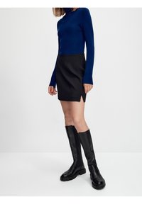 Maglione a collo alto lungo blu navy, gonna mini nera con spacchi laterali e stivali neri in pelle alti fino al ginocchio con suole in gomma.