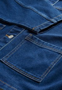 Jachetă albastră din denim cu buzunar la piept vizibil, nasture și cusături portocalii contrastante pe material pliat.