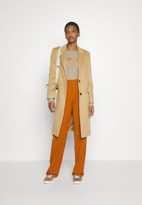 Cappotto beige lungo su una maglietta a coste verde, abbinato a pantaloni arancioni a gamba larga. Sneakers marroni con dettagli in pizzo. Borsa semplice.