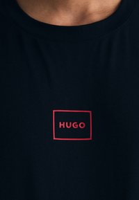 Tricou negru din bumbac, cu guler rotund și un logo rectangular roșu "HUGO" centrat pe față. Textură netedă, design casual.
