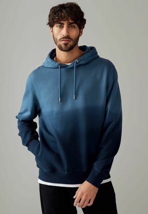 Blauwe gradient hoodie met een trekkoord, met een zachte textuur, geribbelde manchetten en een band onderaan. Gelaagd over een wit shirt.