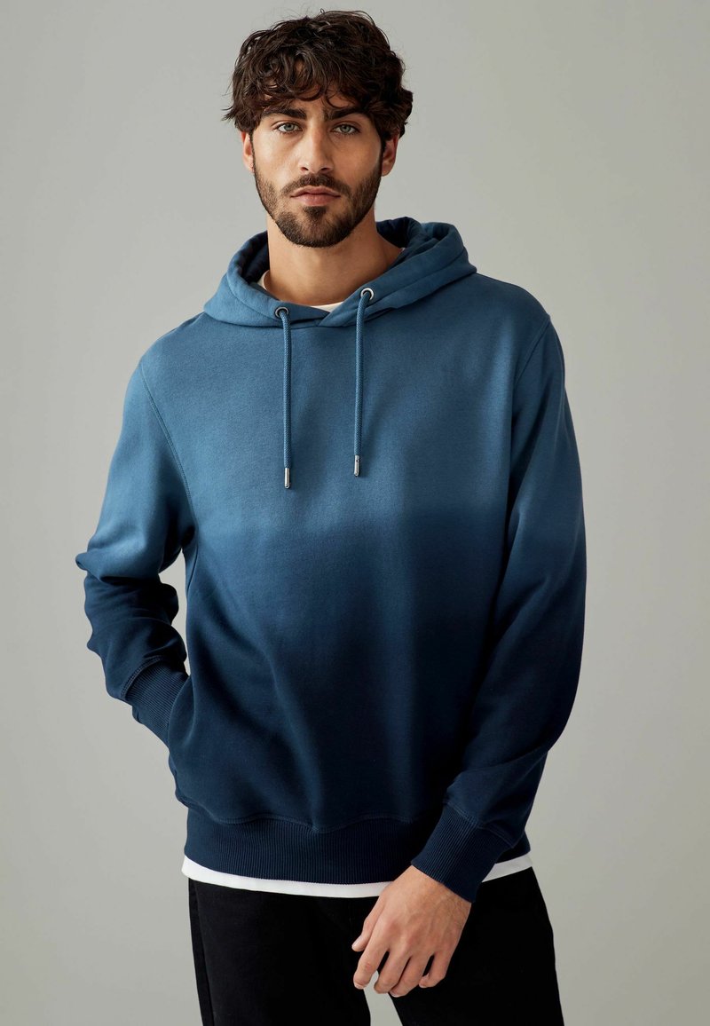 Blauwe gradient hoodie met een trekkoord, met een zachte textuur, geribbelde manchetten en een band onderaan. Gelaagd over een wit shirt.