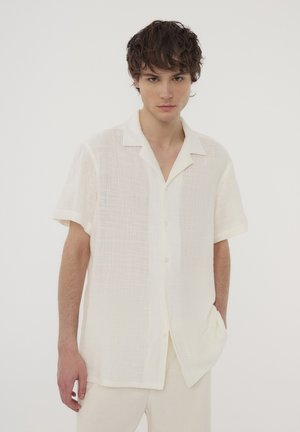 Camicia a maniche corte con colletto di un beige chiaro, realizzata in tessuto testurizzato. Presenta chiusura con bottoni e una vestibilità rilassata. Indossata con pantaloni abbinati.