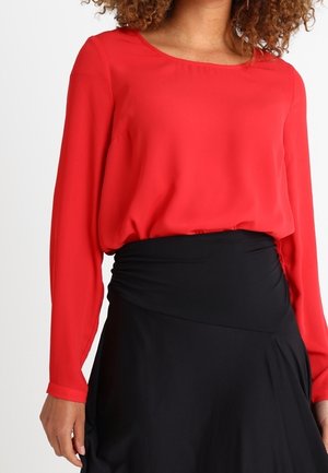 Femme portant un chemisier rouge à manches longues, rentré dans une jupe noire taille haute avec des détails froncés à la taille.