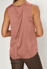 Kvinna klädd i ärmlös topp i dämpad rosafärg och mörkbruna byxor, sedd bakifrån mot en neutral bakgrund.