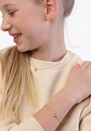 Een goudverguld sieradenset met hartvormige oorbellen, een halsketting en een armband met subtiele details, gedragen tegen een lichtbeige trui.