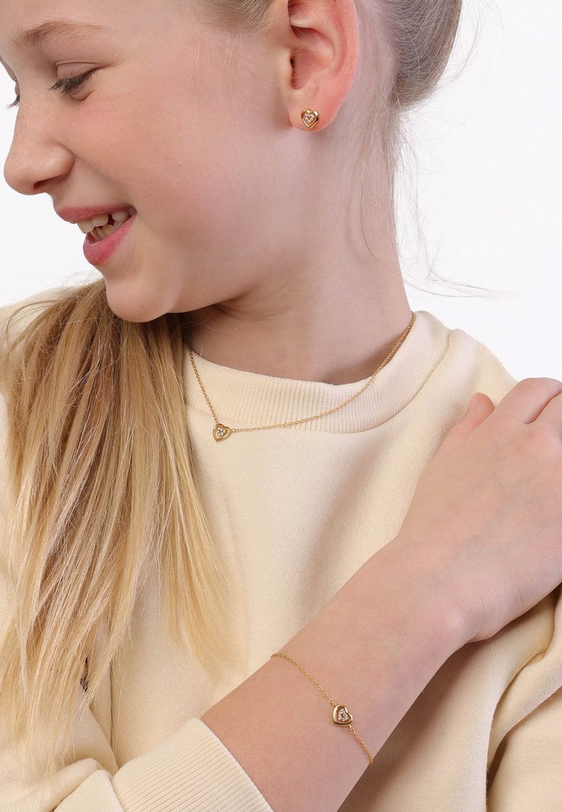 Een goudverguld sieradenset met hartvormige oorbellen, een halsketting en een armband met subtiele details, gedragen tegen een lichtbeige trui.