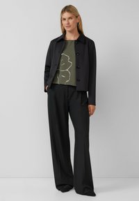 Zwarte getailleerde jas met ronde kraag, front knopen, groene top met bloemmotief en zwarte wijde broek. Glad materiaal, minimale details.