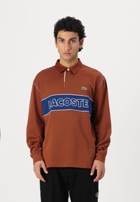 Hnedá polokošeľa s dlhým rukávom a námorníckym horizontálnym pruhom, na ktorom je logo "LACOSTE". Vyrobená z bavlny s bielymi detailami a golierom.