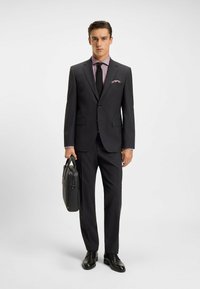 Costume noir sur mesure avec une veste ajustée, chemise blanche à motif à carreaux violets et cravate noire. Tient une mallette en cuir noir. Chaussures noires.