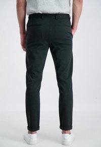 Pantaloni su misura verde scuro con una texture liscia, fit slim e orli arrotolati, abbinati a sneaker bianche. Due tasche posteriori visibili.