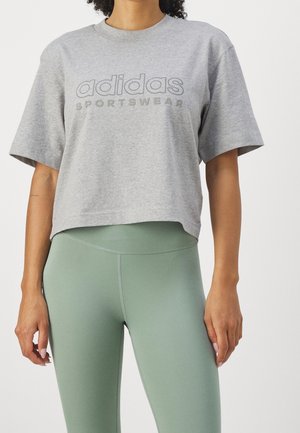 Mujer vistiendo una camiseta corta Adidas Sportswear de color gris claro y mallas de talle alto en verde salvia, de pie frente a un fondo liso.