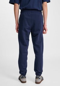 Pantaloni della tuta blu navy con una vita elastica, gambe affusolate e una texture morbida, abbinati a sneaker grigie con dettagli blu.
