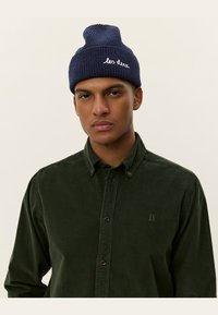 Navy knitted beanie with a rolled cuff and white script text; paired with a dark green corduroy button-up shirt featuring subtle stitching.