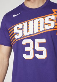 Paarse katoenen T-shirt met de tekst "Suns" in grote witte letters, oranje accenten en het nummer "35" in witte perforatiestijl. Nike-logo op de schouder.