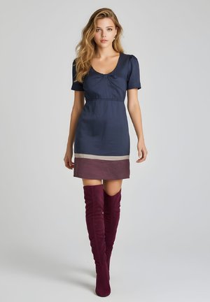 Mujer con cabello largo y rubio que lleva un vestido corto azul marino y burdeos y botas burdeos hasta el muslo, de pie sobre un fondo gris claro.