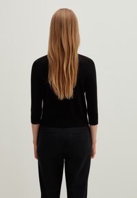 Černý přiléhavý svetr s tříčtvrtečními rukávy, vyrobený z lehkého materiálu. Nošen spolu s tmavými kalhotami. Zezadu jsou vidět dlouhé vlasy.