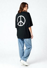 Svart överdimensionerad t-shirt med en stor vit fredssymbol på baksidan. Bärs med ljusblå baggy jeans och vita sneakers.