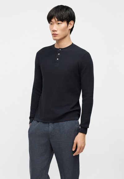 Άνδρας που φοράει μαύρο μπλουζάκι henley με μακριά μανίκια και γκρι παντελόνι, στέκεται με τα χέρια εν μέρει στις τσέπες, μπροστά σε απλό φόντο.