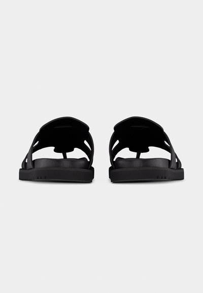Ζεύγος μαύρων πλατύφθαλμων σανδαλιών με ανοιχτή πλάτη και φαρδιές λωρίδες, εμφανιζόμενα από πίσω σε απλό φωτεινό φόντο.