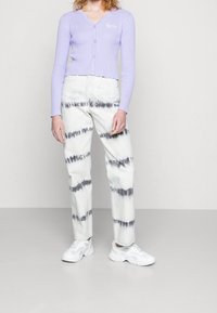 Lila röyhelöinen neuletakki v-kaula-aukkolla ja napeilla, yhdistettynä valkoisiin tie-dye-farkkuihin, joissa mustia kuvioita, viimeistelty valkoisilla lenkkareilla.