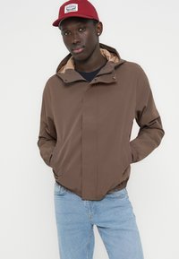 Καφέ μπουφάν με κουκούλα και φερμουάρ μπροστά, ελαστικές μανσέτες και μπεζ εσωτερικό, συνδυασμένο με μπλε τζιν και κόκκινο καπέλο.