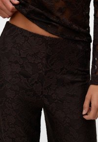 Gros plan d'une personne portant un pantalon en dentelle florale noire et un haut en dentelle assorti, une main ajustant la taille, laissant apparaître la peau à travers le tissu.