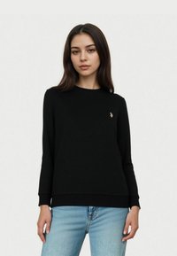 Czarny sweter z długim rękawem, z prążkowanymi mankietami i dołem, z małym złotym logo na lewej piersi. Połączony z jasnoniebieskimi dżinsami.