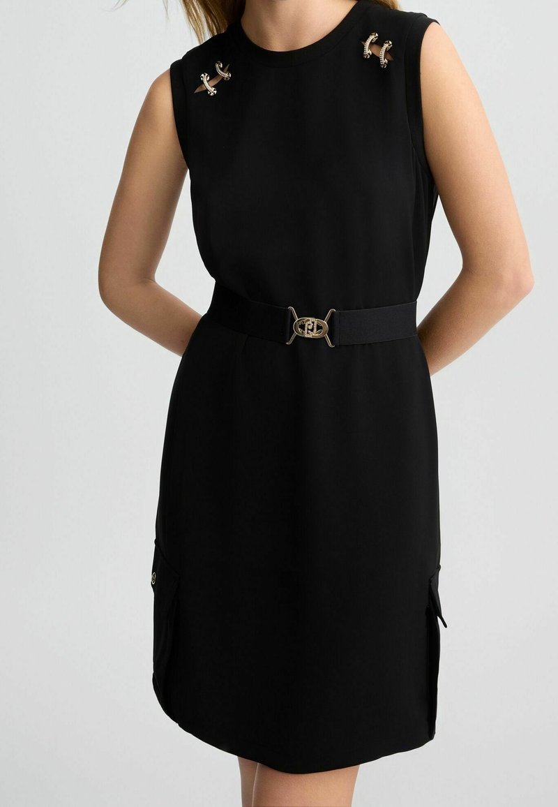 Robe noire sans manches avec ceinture à boucle dorée, épingles décoratives dorées sur les épaules et poches latérales, portée par une personne les bras dans le dos.