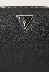 Portafoglio in pelle nera testurizzata con un accento triangolare in oro dal logo Guess a forma di punto interrogativo, chiuso da una zip.