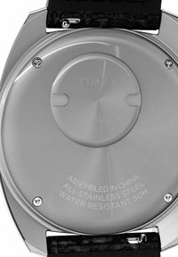 Fond de montre en acier inoxydable avec texte gravé. Présente une forme ronde, une texture métallique lisse et un attachement de bracelet noir.