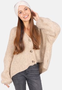 Gilet en tricot beige avec de grandes manches, agrémenté de trois boutons décoratifs. Associé à un jean foncé et un bandeau clair.